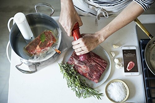 Person preparing meat for sous vide cooking with herbs and smartphone nearby.